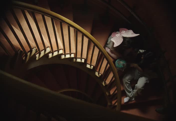 Two individuals are descending a spiral staircase with the one in front carrying a blue globe the staircase features wooden steps and a wooden handrail illuminated by lights embedded within the structure casting a warm glow