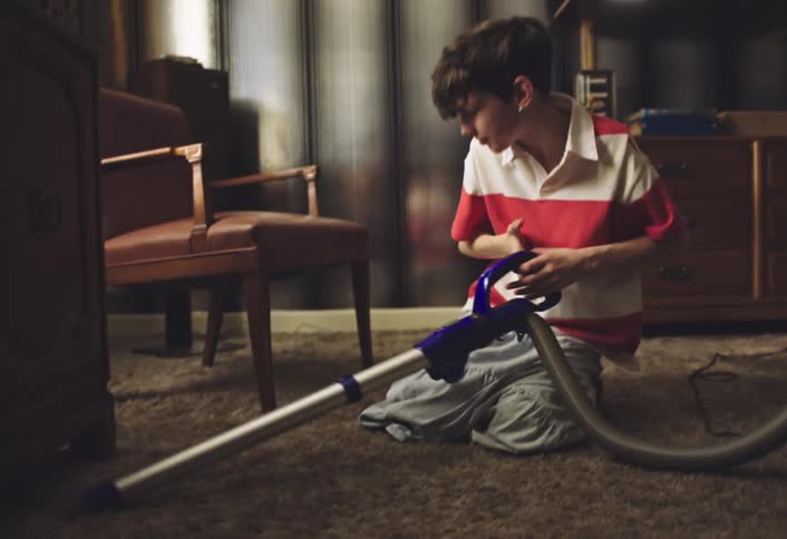 A child wearing a red and white striped polo shirt and lightcolored pants kneeling on a carpeted floor while operating a blue and gray vacuum cleaner the room appears to be a bedroom with patterned curtains and wooden furniture in the background