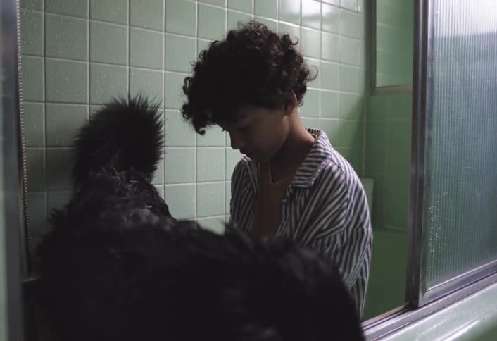 A person with dark curly hair is seen in a bathroom standing in front of a tub with light green tiled walls they are bathing a large black furry dog gently scrubbing its back