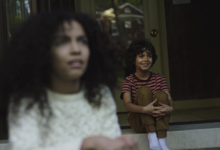 Curly haired girl in the foreground watching a boy in the background with a happy expression talking