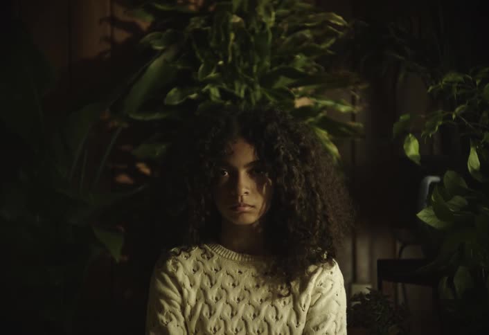 A woman in a white knitted sweater and light green pants sits crosslegged on the floor surrounded by lush potted houseplants