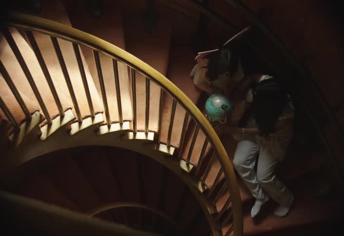 Two children are seen descending a winding wooden staircase with one child carrying a blue globe the staircase is warmly lit by lights positioned within the railing creating a cozy and inviting atmosphere