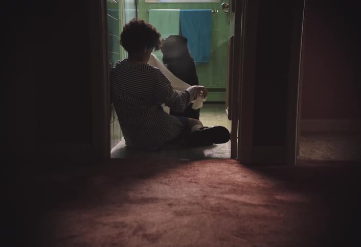 A darkhaired person in a striped shirt is seen on the floor of a dim room partially obscured by a doorway they are interacting with a black dog eventually wrapping it in a white towel