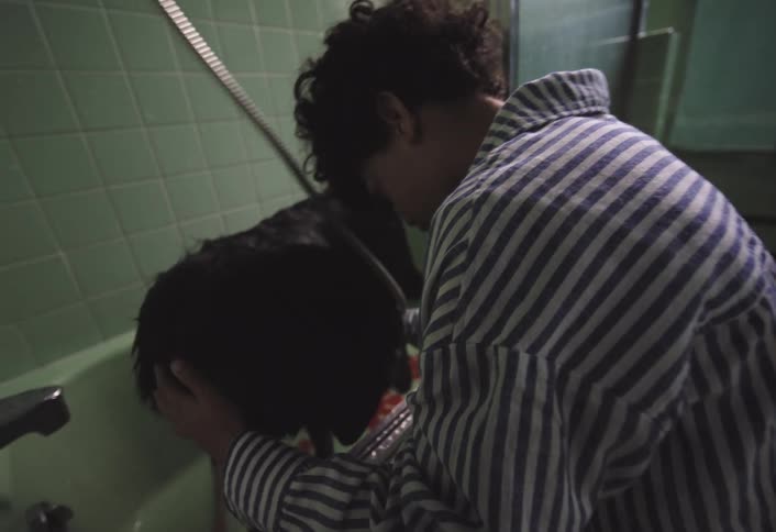 A child with curly hair and a striped shirt is seen bathing a black dog in a tub with light green tiled walls they are using a handheld shower head to rinse the dogs fur focusing intently on the task