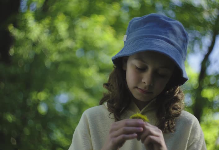 A young person wearing a blue bucket hat and a lightcolored longsleeved shirt is shown outdoors likely in a park or forest with blurred green foliage in the background they are holding and examining a small yellow flower with a focused expression