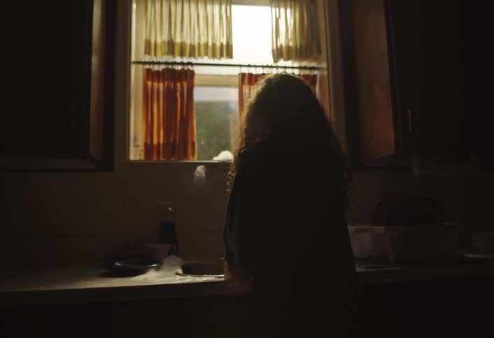 A young person with long dark curly hair stands silhouetted against a window in a dimly lit room facing away from the camera the window has two sets of curtains one lighter and one darker orange through which some light is visible