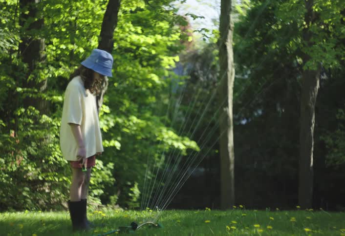 The girl stands in front of a sprinkler on the lawn