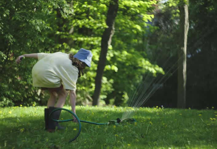 A child in a yellow top and blue bucket hat is standing in a green grassy area with a sprinkler spraying water they walk over to the sprinkler and bend down to adjust it
