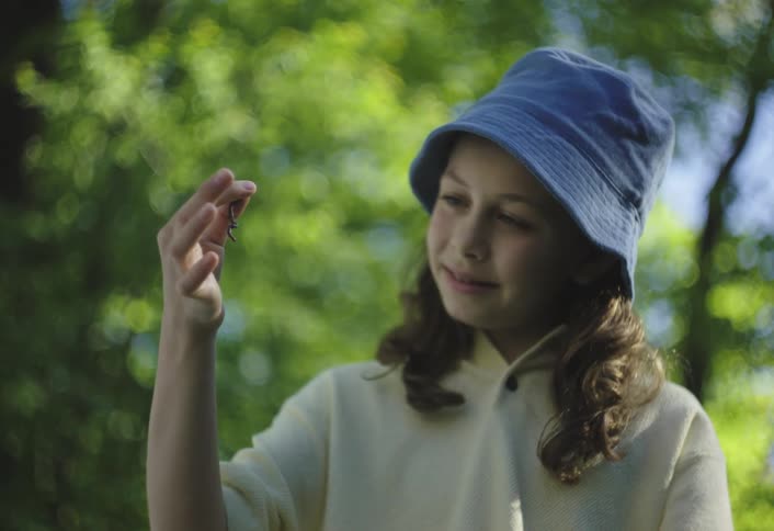 A girl stands outside holding a worm between her fingers while smiling at it