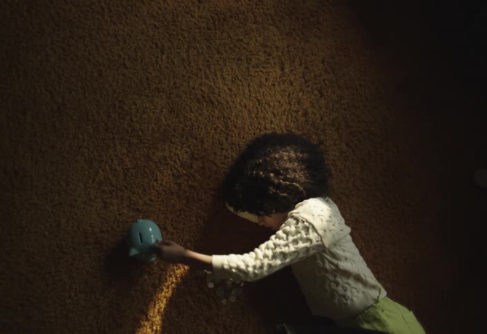 Girl lying on the carpeted floor puts coins into her piggy bank before turning to look upward