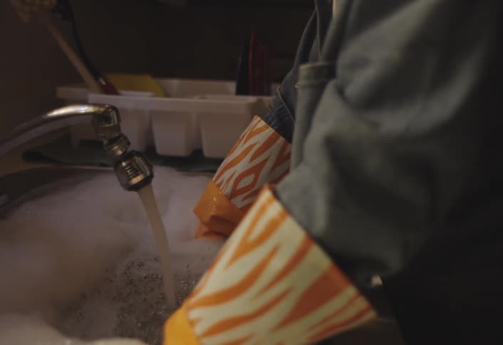 A person wearing orange cleaning gloves is seen washing a dark blue plate in a sink filled with soapy water the water runs from the faucet into the sudsy sink creating bubbles as they clean