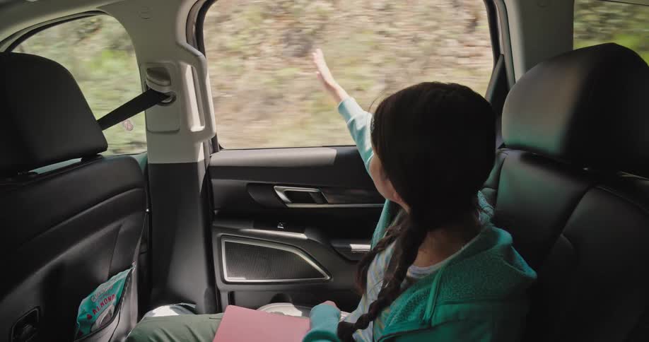 A young girl with braided hair sitting in the back seat of a car she is looking out the window and waving her hand seemingly enjoying the view as the car moves