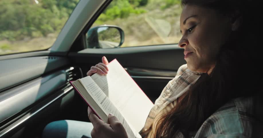 A woman in the front passenger seat of a car is reading a book with a smile on her face at midday