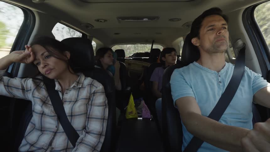 A family of four in a car with the father driving and the mother in the passenger seat both appearing tired or bored two children are visible in the back seat