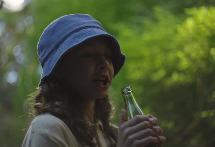 A child wearing a hat is standing outdoors in the woods at midday sipping soda from a bottle