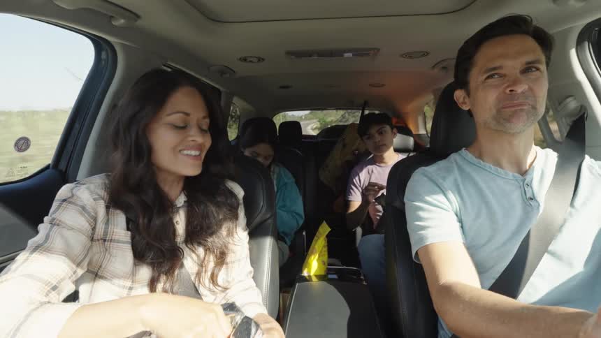 A family is seen inside a car with a man driving and a woman in the passenger seat engaging in conversation several children are visible in the back seats suggesting a road trip or family outing