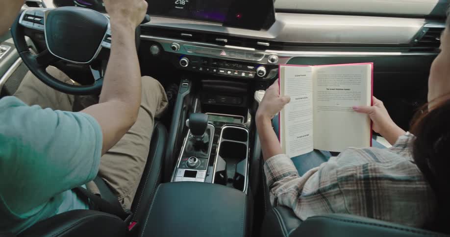 Female passenger pauses in reading a book to turn the radio back on after the driver turned it off