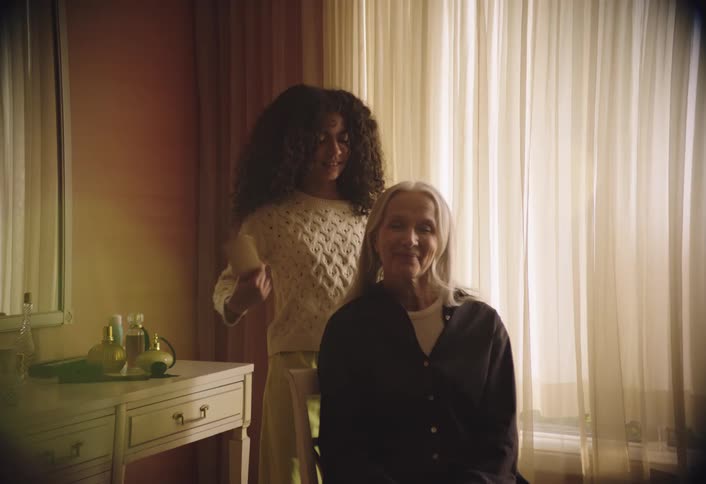A young woman with long curly hair is seen standing behind an older woman with long gray hair who is seated in front of a window the young woman is gently combing or brushing the older womans hair who appears to be enjoying the interaction