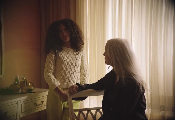 This video features two women one with dark curly hair and the other with long blonde hair interacting in a room with a window providing natural light the darkerhaired woman stands while the blondehaired woman is seated