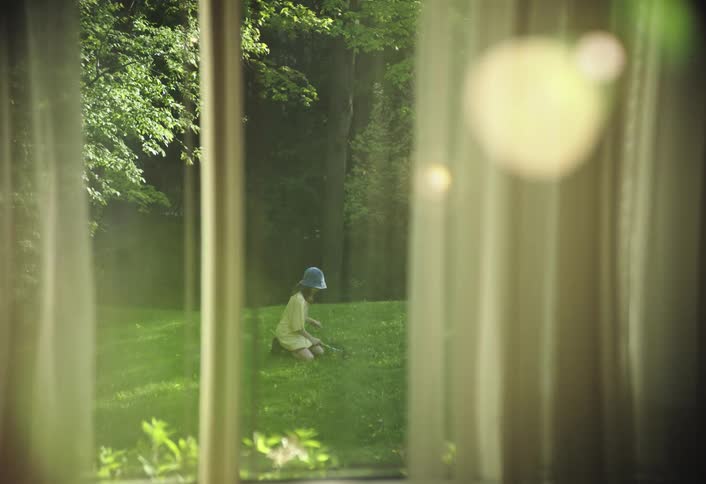 The video shows a view from inside a room looking out through a window with sheer curtains outside a person in a blue hat and lightcolored top is seen kneeling on a bright green lawn seemingly tending to plants or grass