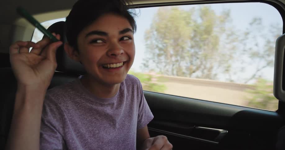 A person with dark hair is seen seated in the back of a car looking out the window with a curious expression the car appears to be in motion as trees and a clear sky are visible through the window