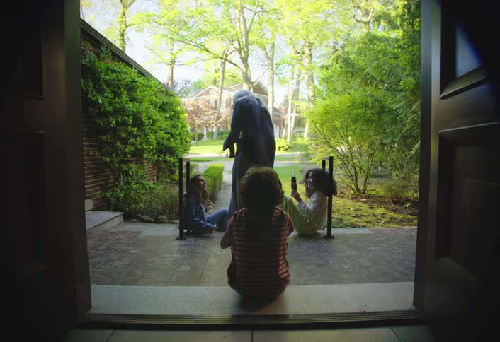 The video shows a group of children and adults gathered outside a house viewed from the dark interior through a doorway the scene is bright and verdant with lush green trees and foliage surrounding the pathway where the individuals are interacting