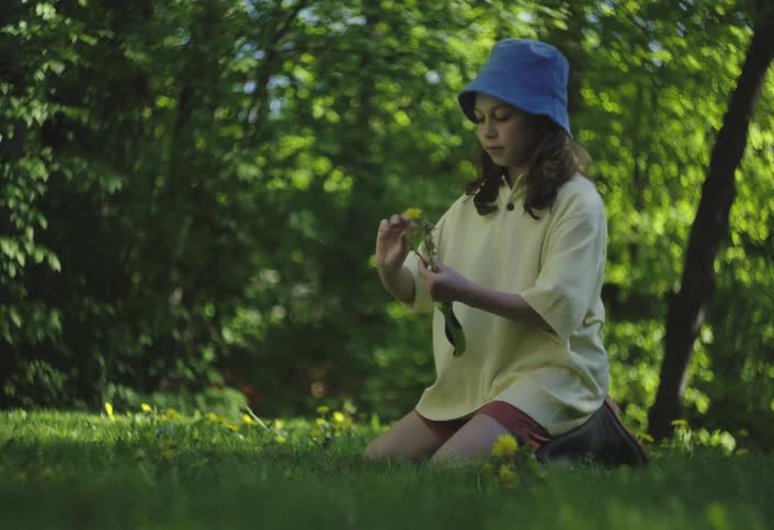 A person wearing a yellow top and a blue bucket hat is kneeling on a lush green lawn surrounded by yellow flowers they are attentively examining a yellow flower they hold in their hand then place it back down