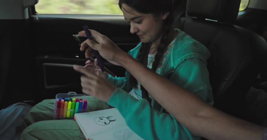 A girl sits in a car drawing in her notebook with a container of colorful markers on her lap while the person beside her playfully snatches a pen sparking a lighthearted tussle