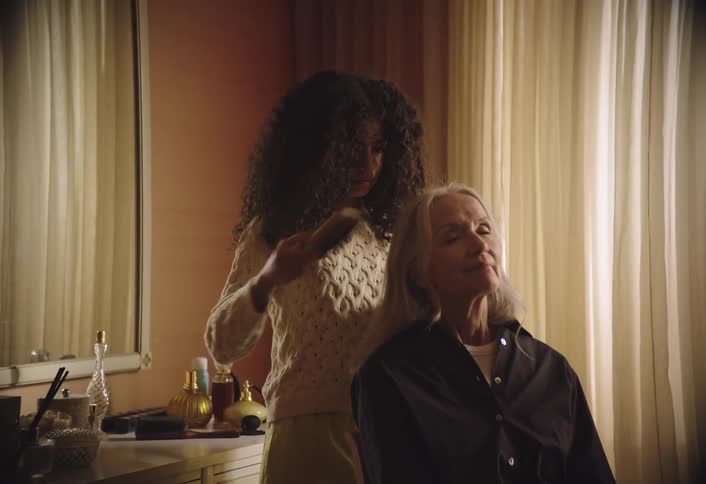 A teenage girl gently styles an elderly womans hair beside a vanity table cluttered with grooming products in a softly lit room with sheer curtains
