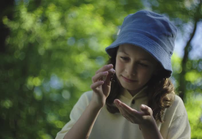 A girl in a blue hat holds a small worm close to her face with a curious expression
