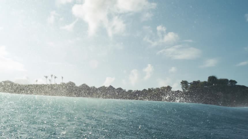Waves crash hard against a rocky shore under a blue sky with scattered clouds and vegetation in the background