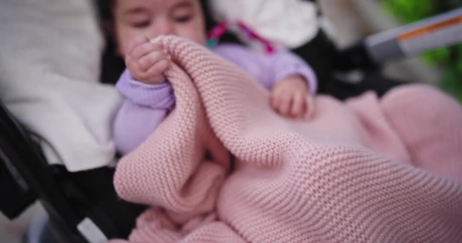 Baby sits in a stroller with a knit blanket on her lap near a curb
