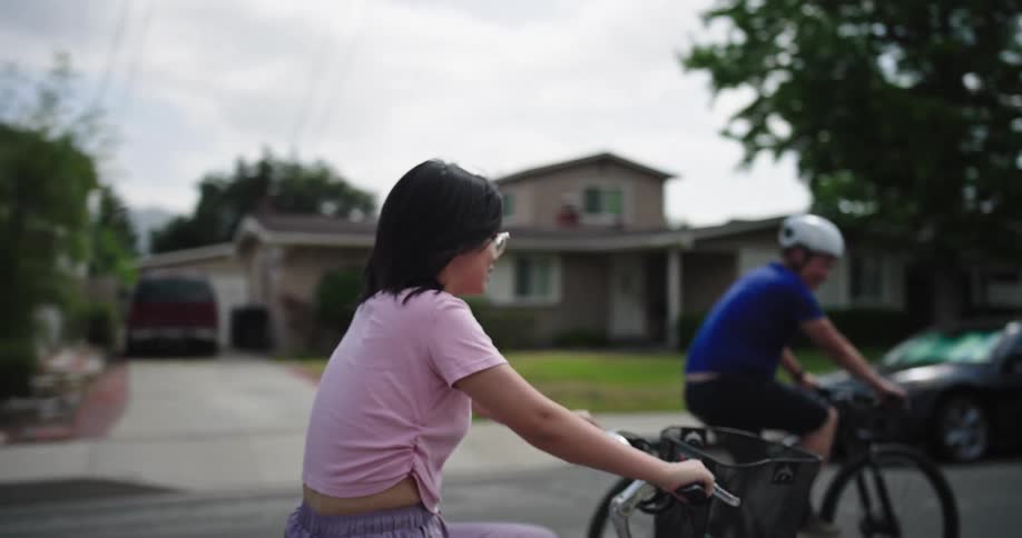 A young woman with dark hair and glasses wearing a light pink tshirt and lightcolored shorts is seen riding a bicycle she is smiling and appears to be enjoying herself in the background another person a man wearing a blue shirt and a white helmet