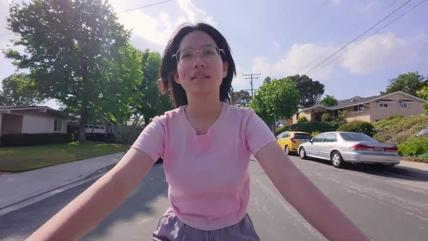 A firstperson perspective shows a woman with dark hair and glasses riding a bicycle looking forward with a slight smile she is wearing a pink tshirt and light purple shorts the setting appears to be a suburban street with houses trees and parked