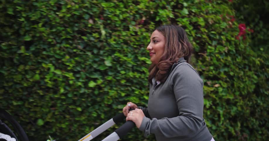 Mother pushes her baby in a stroller walking along the sidewalk past tall bushes