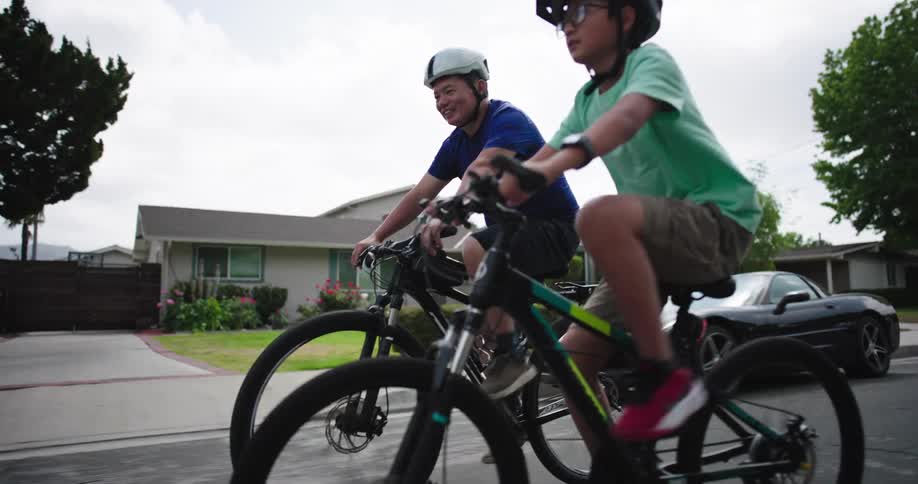Two males are seen cycling on a residential street the person in the foreground wearing a light green tshirt and shorts with red and white shoes is on a black and green bicycle the person behind him is wearing a blue tshirt and black shorts