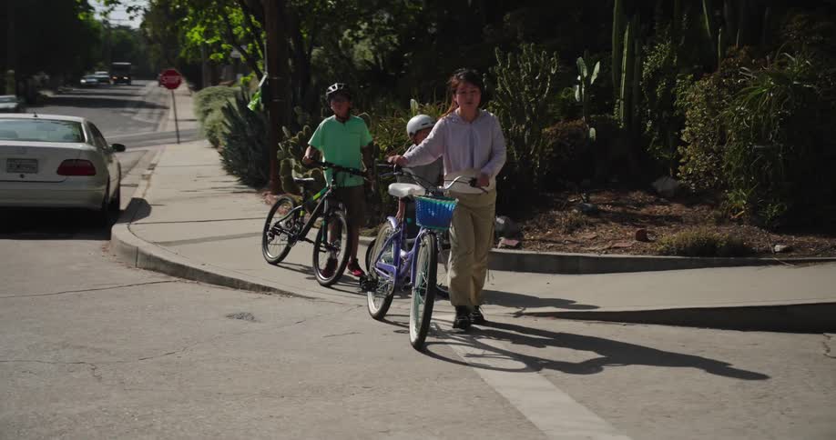 Two boys and a teenage girl walk their bicycles across the street in a quiet neighborhood