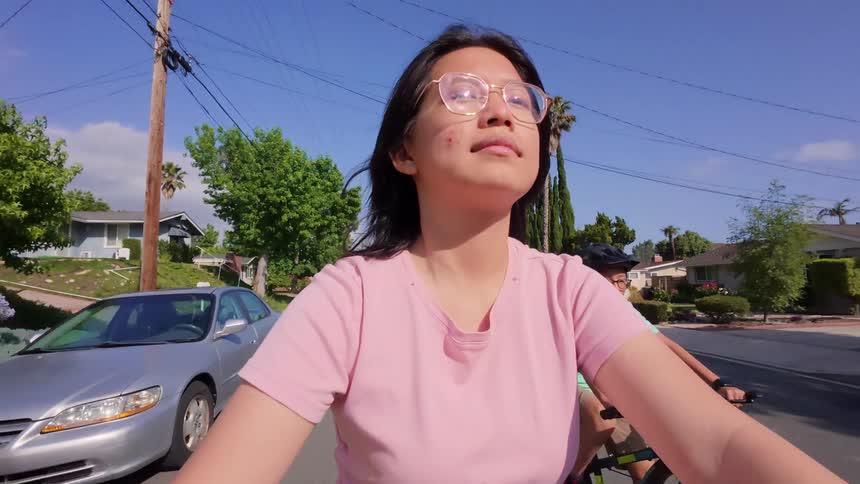 Two individuals are cycling along a residential street on a clear sunny day the person in the foreground wearing a pink shirt and glasses is looking directly ahead while the person behind them wearing a green shirt and a helmet