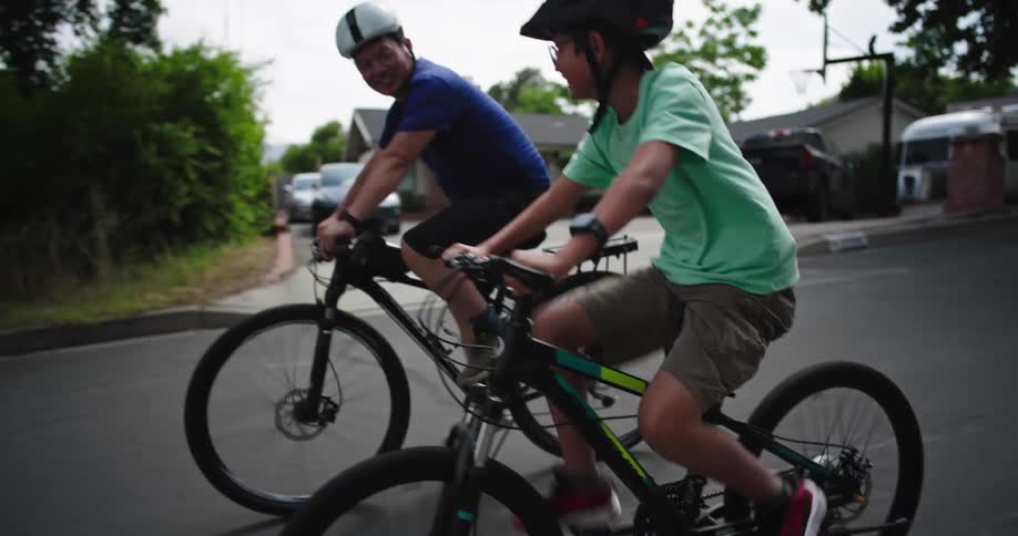 Two male cyclists one adult and one child both wearing helmets are riding bicycles on a paved road the adult is on a black bike with a white helmet and the child is on a multicolored bike with a black helmet actively pedaling
