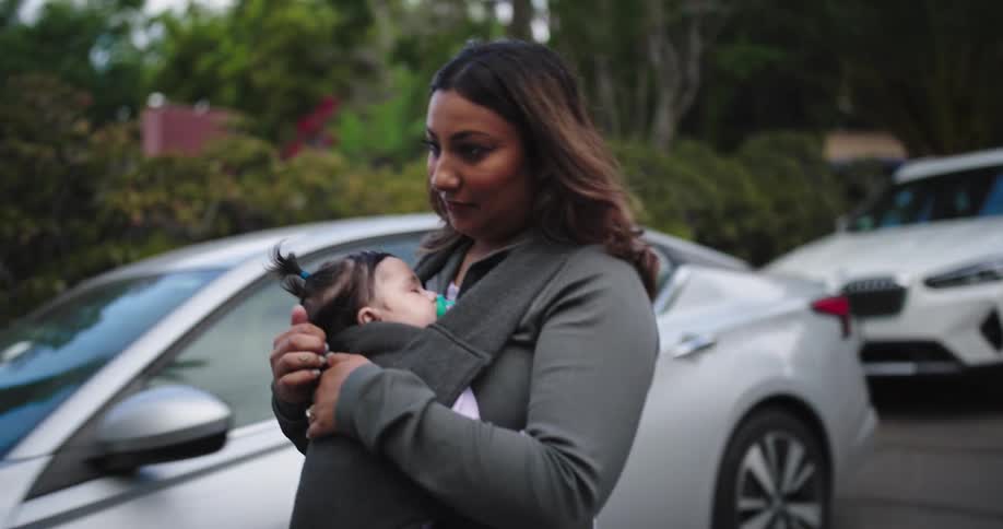 A woman cradles a sleeping baby in a front carrier while standing near cars