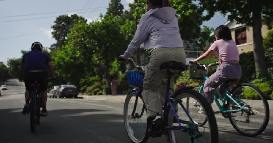 A group of people including adults and children are cycling down a residential street on a sunny day they are all riding away from the camera with the person on the far right wearing a pink shirt and purple pants on a turquoise bicycle