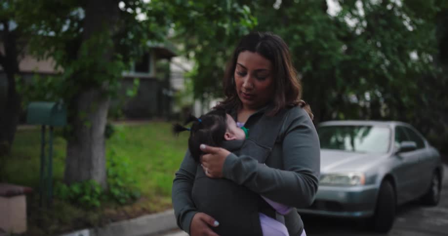 A woman with long dark hair wearing a grey longsleeved top is holding a baby in a black carrier against her chest the baby has dark hair tied in two small ponytails and a green pacifier in its mouth the background features a residential street