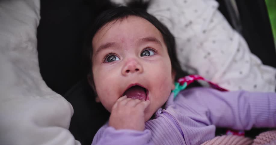 A baby in a purple outfit lying down with fingers in their mouth looking curiously at the camera