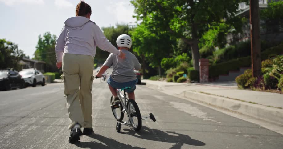 Mother guides her son as the child pedals a bicycle with training wheels down a residential street