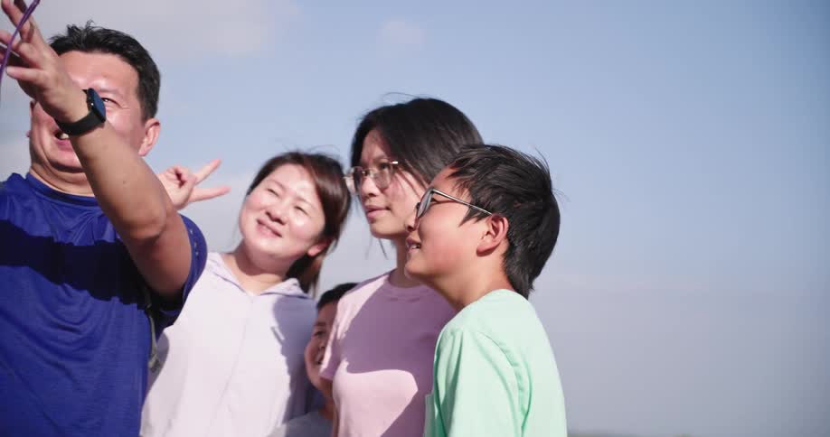 A family of five is posing for a selfie outdoors under a bright sky the father on the left is holding a camera or phone out to capture the photo smiling broadly next to him a woman and three children are also smiling and looking towards the camera