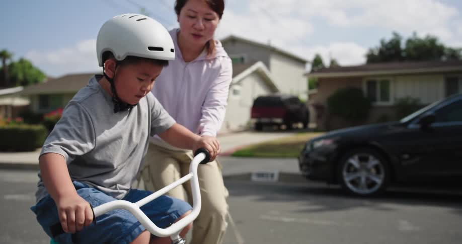 A child wearing a white helmet is learning to ride a bicycle with training wheels on a paved residential street an adult is supporting the child from behind helping them maintain balance and push off