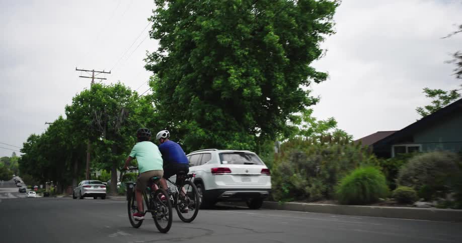 Two individuals wearing helmets are cycling down a residential street flanked by houses and trees the person in the light green shirt and the person in the blue shirt cycle sidebyside moving away from the camera