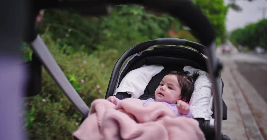 The video shows a closeup of a baby wrapped in a pink blanket lying in a stroller the baby looks around with curiosity as the stroller moves along a sidewalk next to some greenery