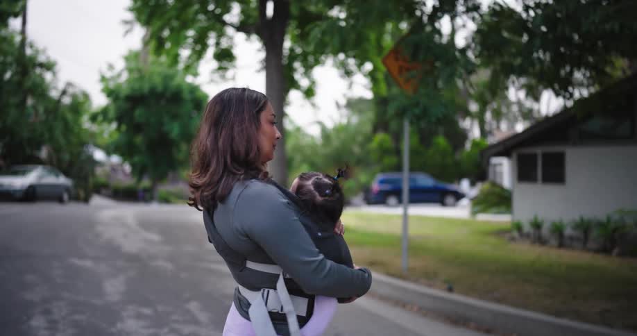 A young woman is carrying her child with her arms wrapped around the baby as she crosses the street