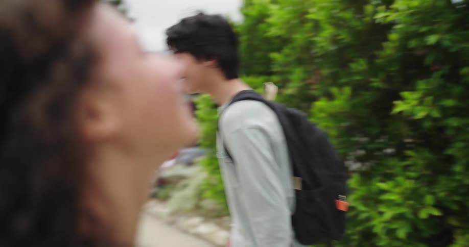 Two young individuals with backpacks are interacting on a sidewalk bordered by green bushes one person has dark hair and is wearing a grey longsleeved shirt while the other has curly hair and is seen from behind with a light blue backpack